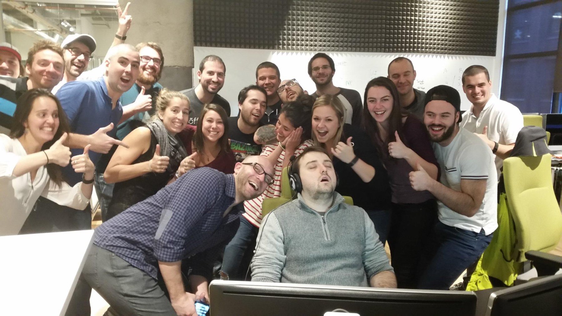 A team crowded around a desk giving a thumbs up, while one person sleeps in the chair.